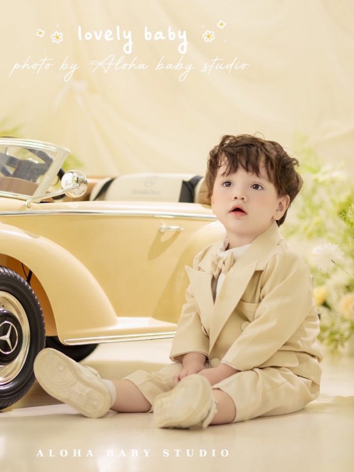 Bé trai mặc vest kem ngồi cạnh xe hơi đồ chơi màu vàng nhạt — concept Baby bé trai tại Aloha.