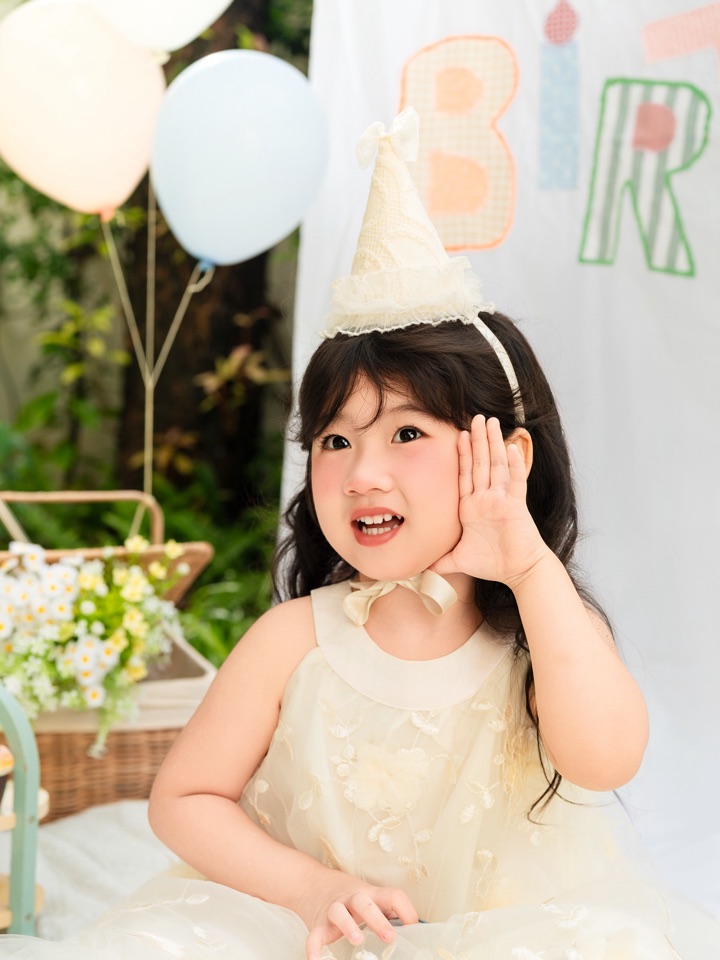 Bé gái mặc váy trắng, đội mũ sinh nhật, tay đưa lên má cười tươi — concept Baby bé gái tại Aloha.