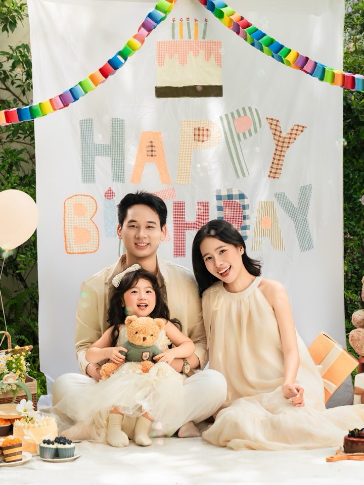 Ba mẹ và bé gái ngồi cùng nhau trước phông Happy Birthday, có bóng bay và gấu bông — concept Family tại Aloha.