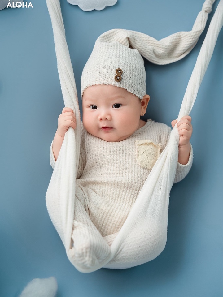 Bé sơ sinh mặc đồ len kem, đội mũ chóp, nằm trong võng vải trên nền xanh nhạt — concept Newborn bé trai tại Aloha.