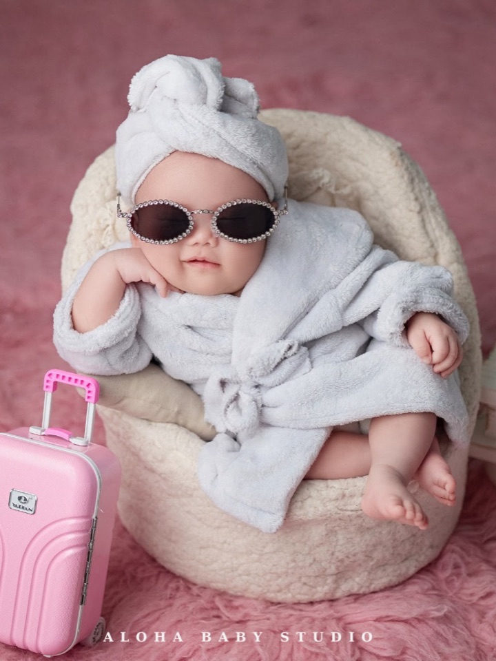 Bé sơ sinh quấn khăn tắm, đội khăn turban và đeo kính râm nhỏ, cạnh vali hồng — concept Newborn bé gái tại Aloha.