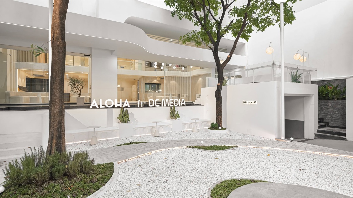 Mặt tiền cơ sở TP.HCM của Aloha: toà nhà trắng có biển ALOHA, sân sỏi trắng, vài cây xanh và ghế đá.