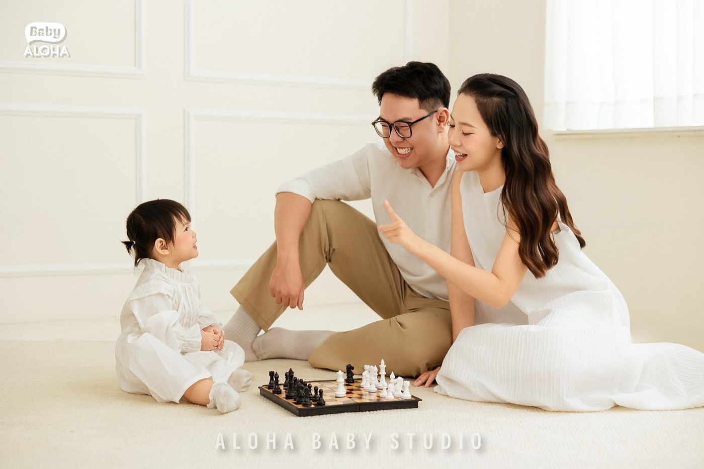 Ba mẹ và bé gái ngồi chơi cờ trên nền kem, cả nhà cùng cười — chụp tại Aloha Baby Studio.
